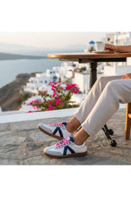 Load image into Gallery viewer, Person wearing white sneakers with blue and pink accents sitting at a table with a scenic background.